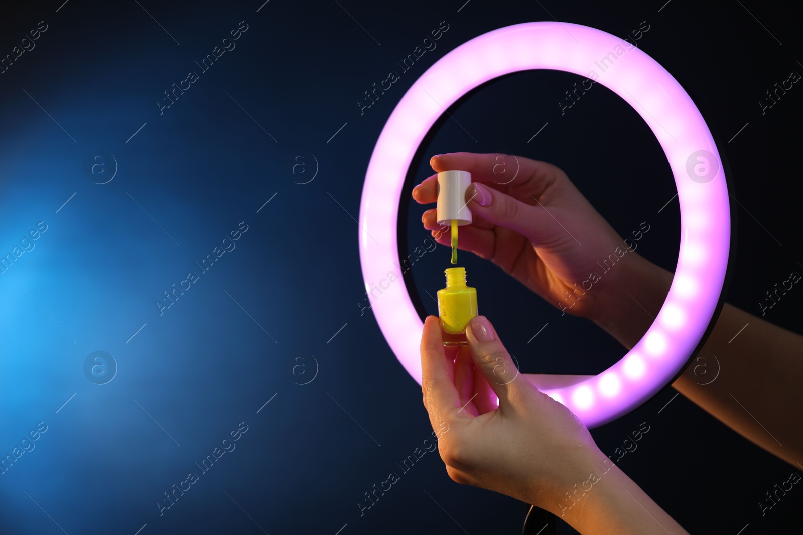 Photo of Woman holding nail polish through ring lamp on dark blue background, closeup. Space for text