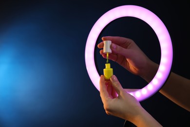 Photo of Woman holding nail polish through ring lamp on dark blue background, closeup. Space for text