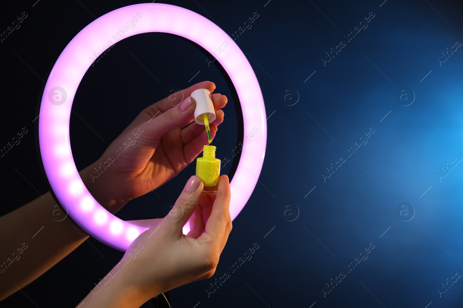 Photo of Woman holding nail polish through ring lamp on dark blue background, closeup. Space for text