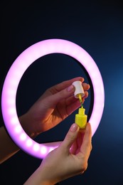 Photo of Woman holding nail polish through ring lamp on dark blue background, closeup