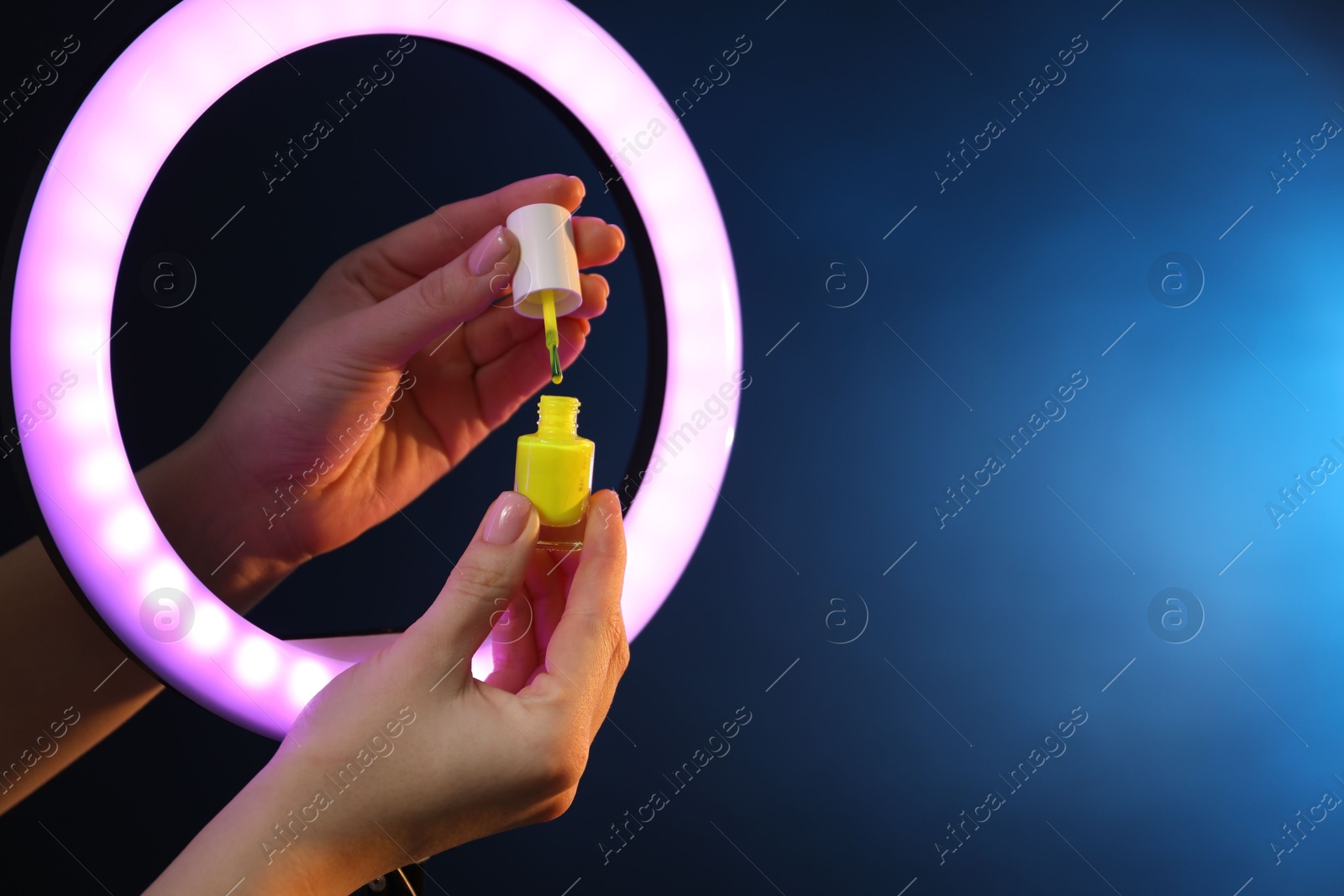 Photo of Woman holding nail polish through ring lamp on dark blue background, closeup. Space for text