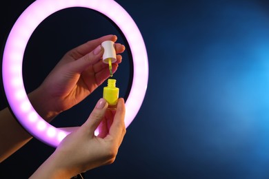 Photo of Woman holding nail polish through ring lamp on dark blue background, closeup. Space for text