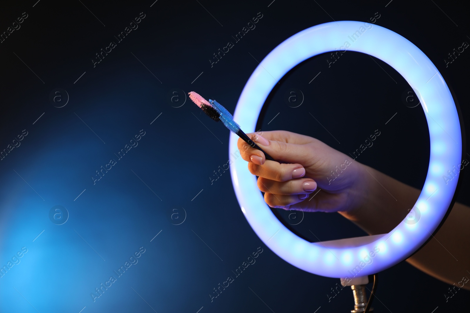 Photo of Woman holding makeup brushes through ring lamp on dark blue background, closeup. Space for text