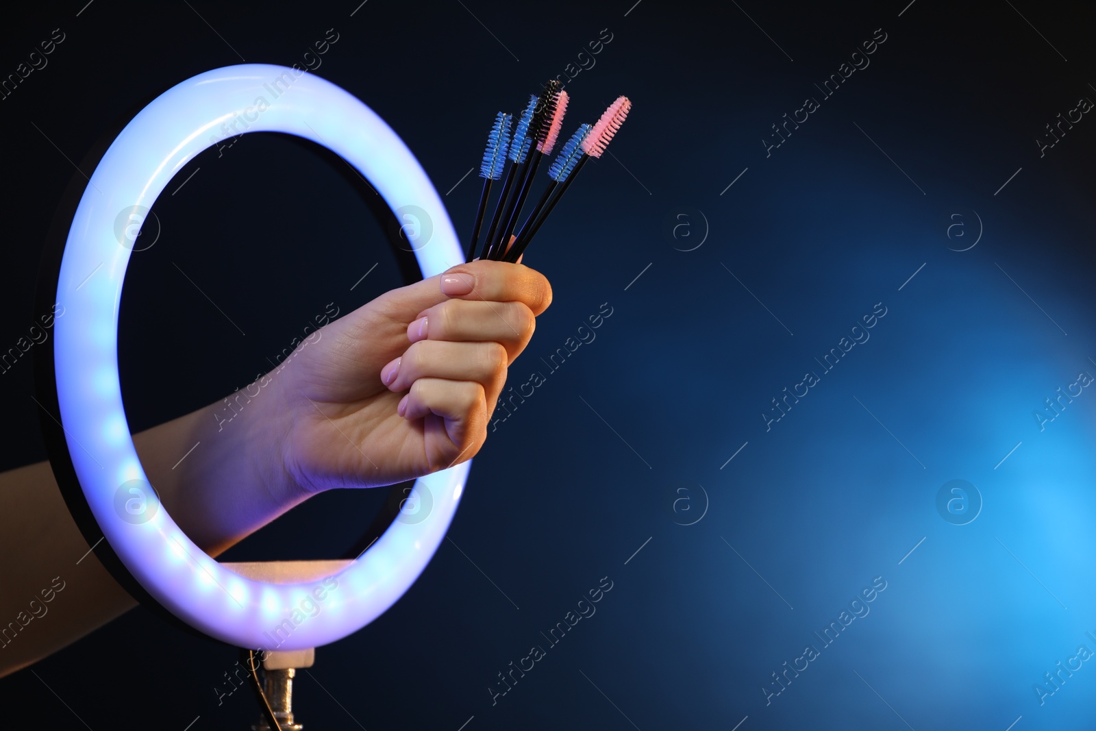 Photo of Woman holding makeup brushes through ring lamp on dark blue background, closeup. Space for text