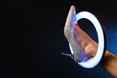 Photo of Woman with ring lamp wearing high heeled shoe on dark blue background, closeup. Space for text