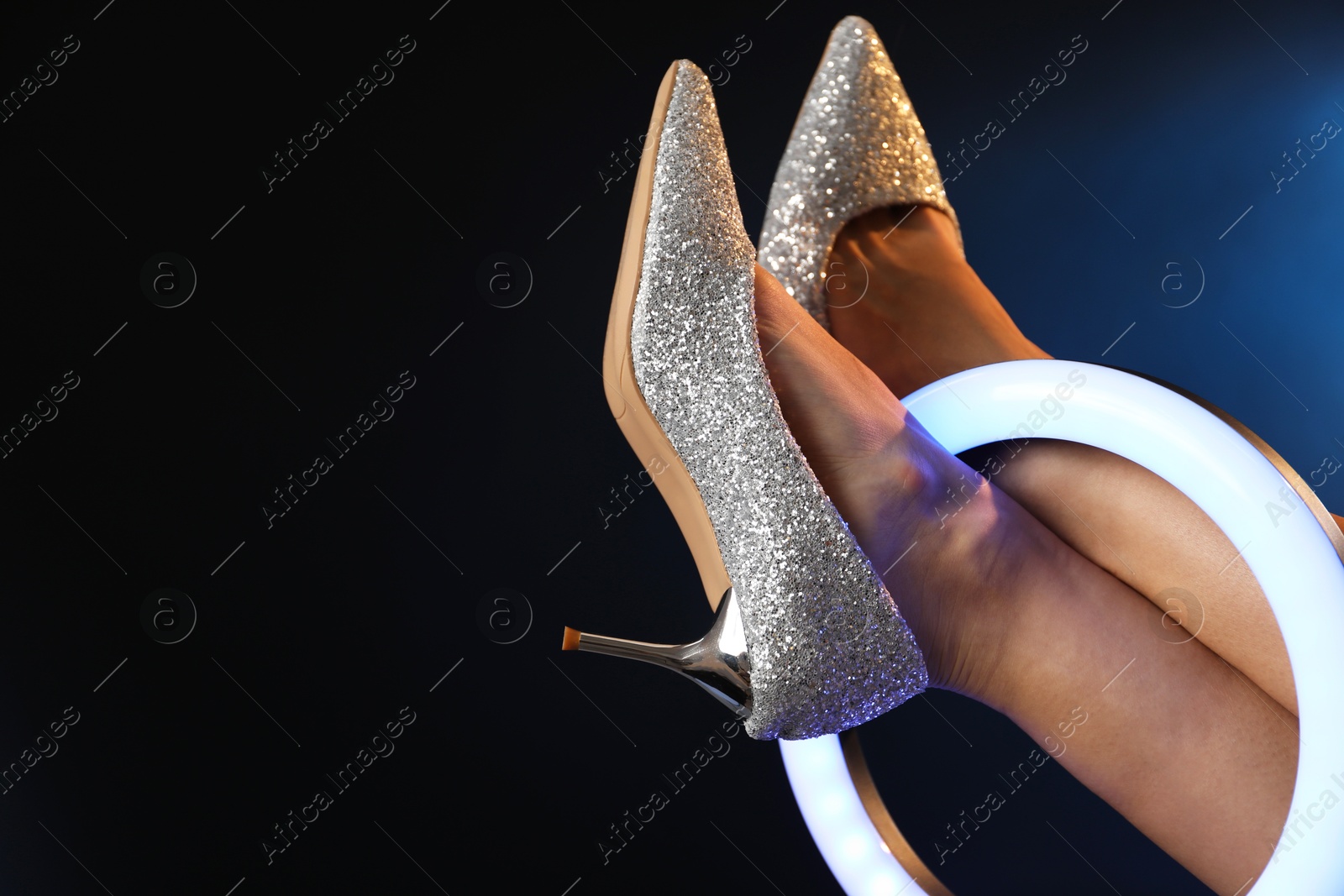 Photo of Woman with ring lamp wearing high heeled shoes on dark blue background, closeup. Space for text