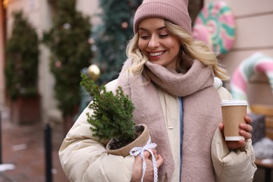 Happy woman with thuja tree and paper cup of coffee on city street, space for text. Christmas season