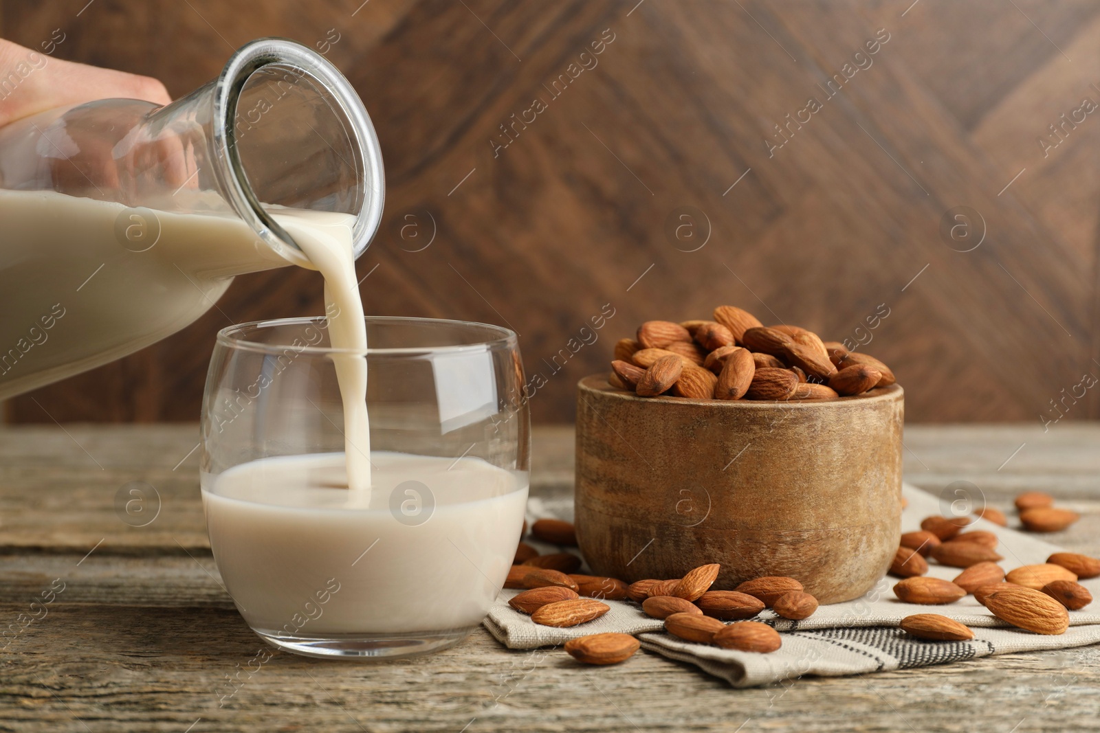 Photo of Woman pouring almond milk into glass and nuts at wooden table, closeup