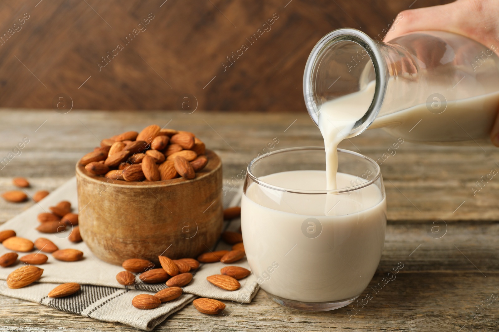 Photo of Woman pouring almond milk into glass and nuts at wooden table, closeup