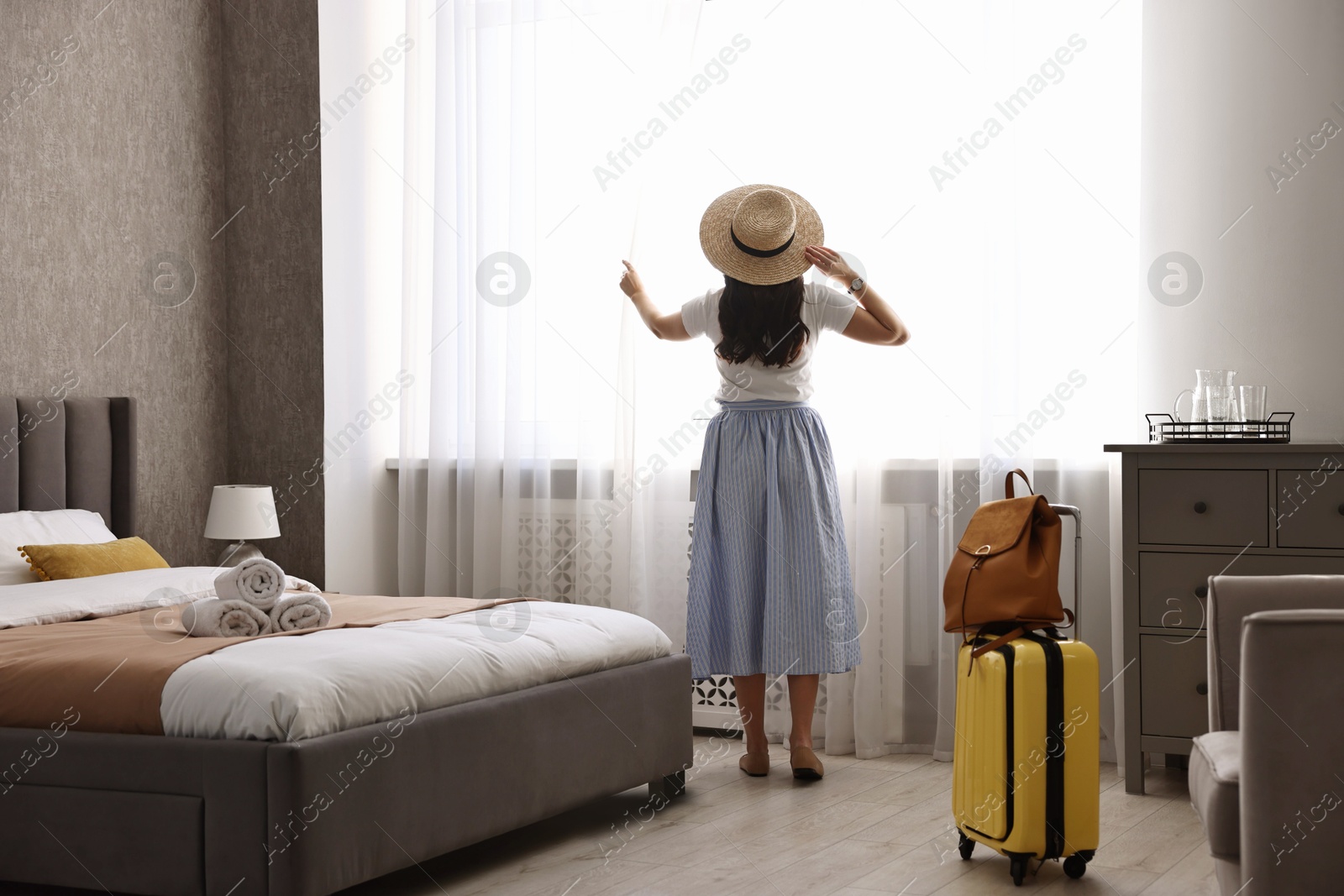 Photo of Traveller with suitcase opening curtains in hotel room, back view