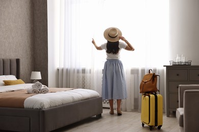 Photo of Traveller with suitcase opening curtains in hotel room, back view