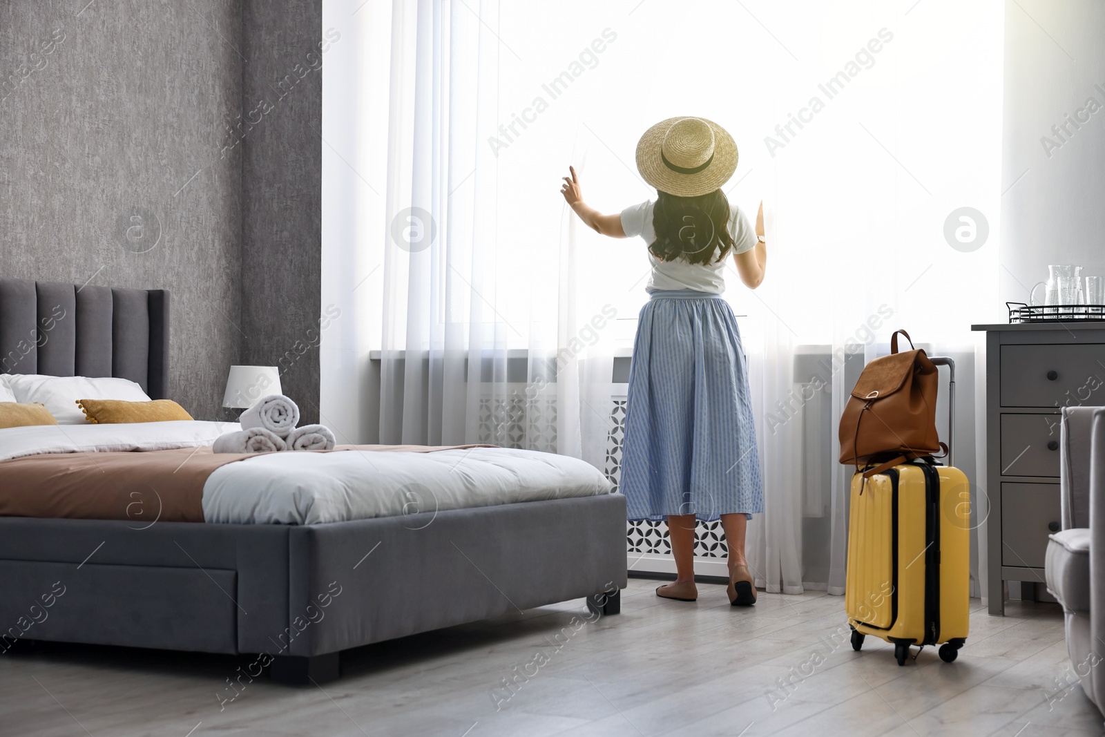 Photo of Traveller with suitcase opening curtains in hotel room, back view