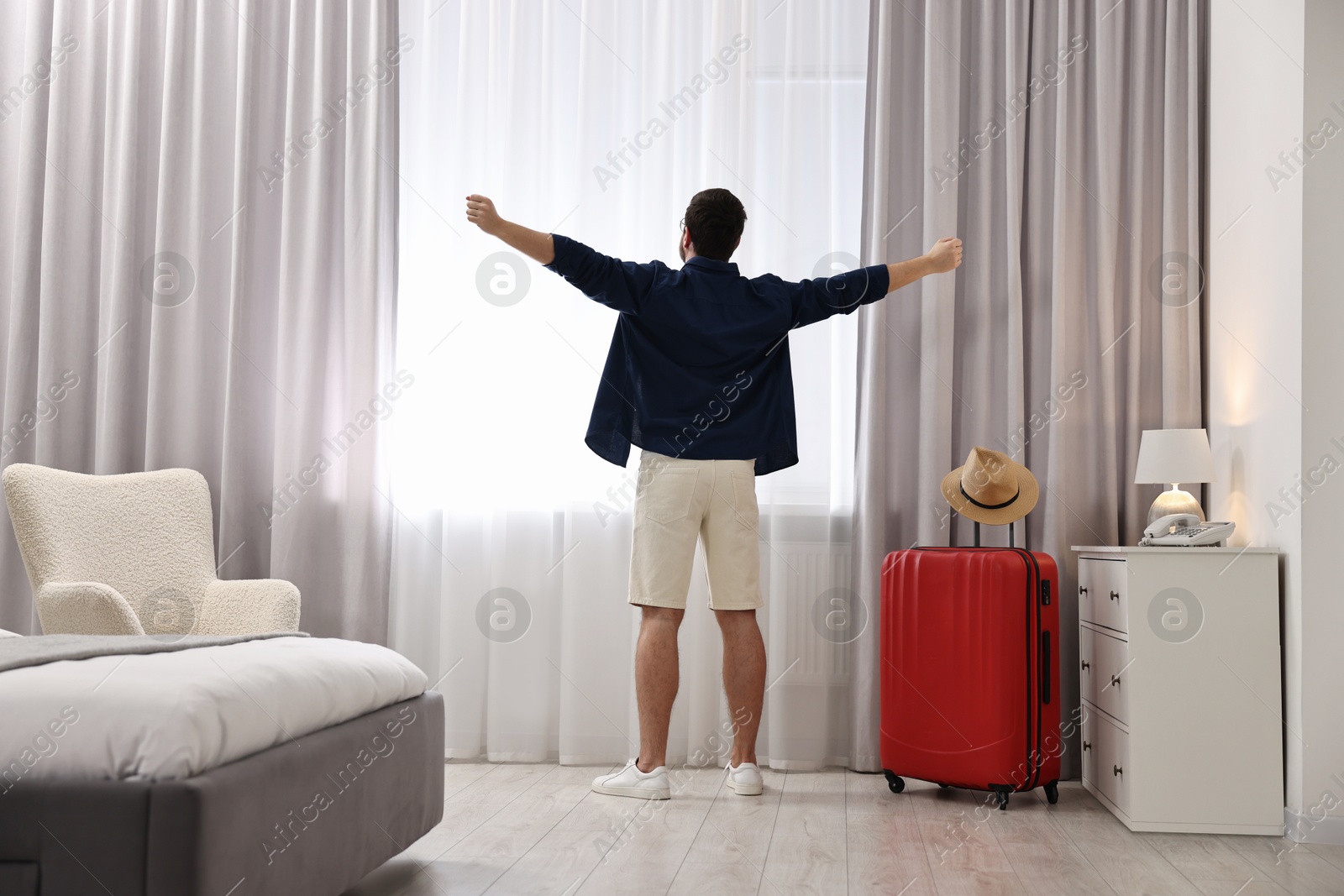 Photo of Traveller and suitcase near window in hotel room, back view