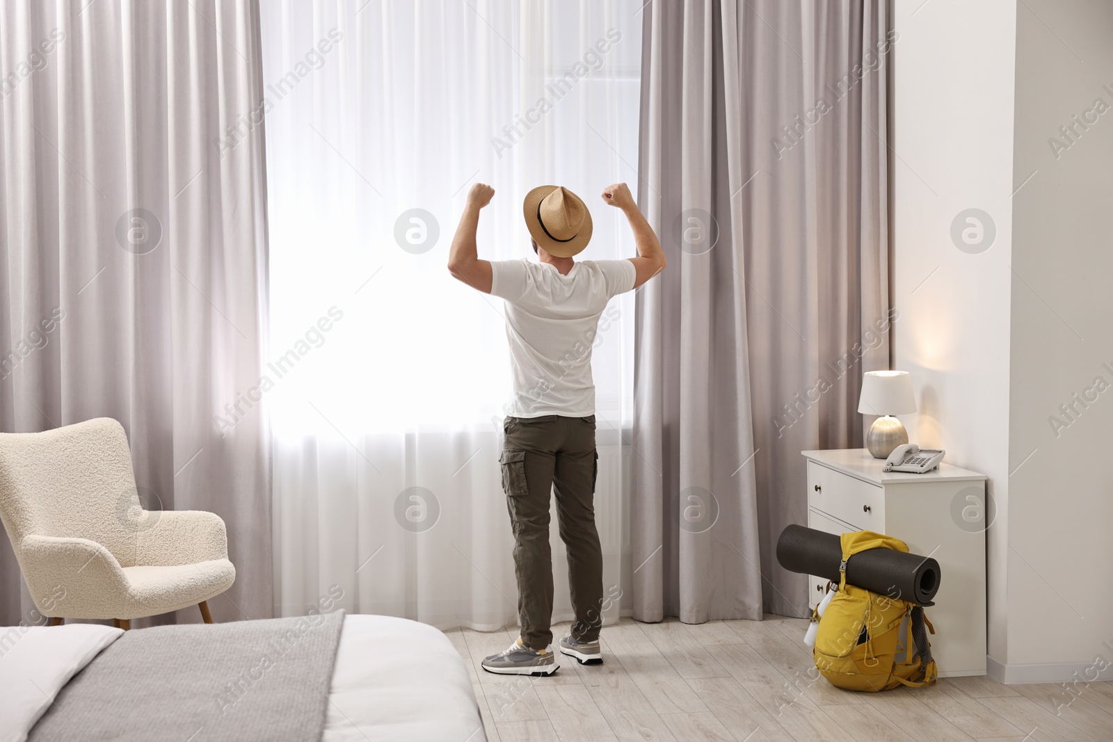 Photo of Traveller with straw hat near window in hotel room, back view