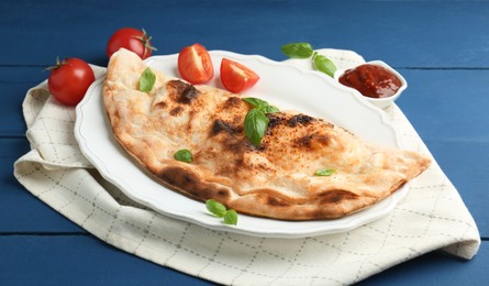 Photo of Tasty calzone with basil, tomatoes and sauce on blue wooden table, closeup