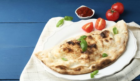 Photo of Tasty calzone with basil, tomatoes and sauce on blue wooden table, above view