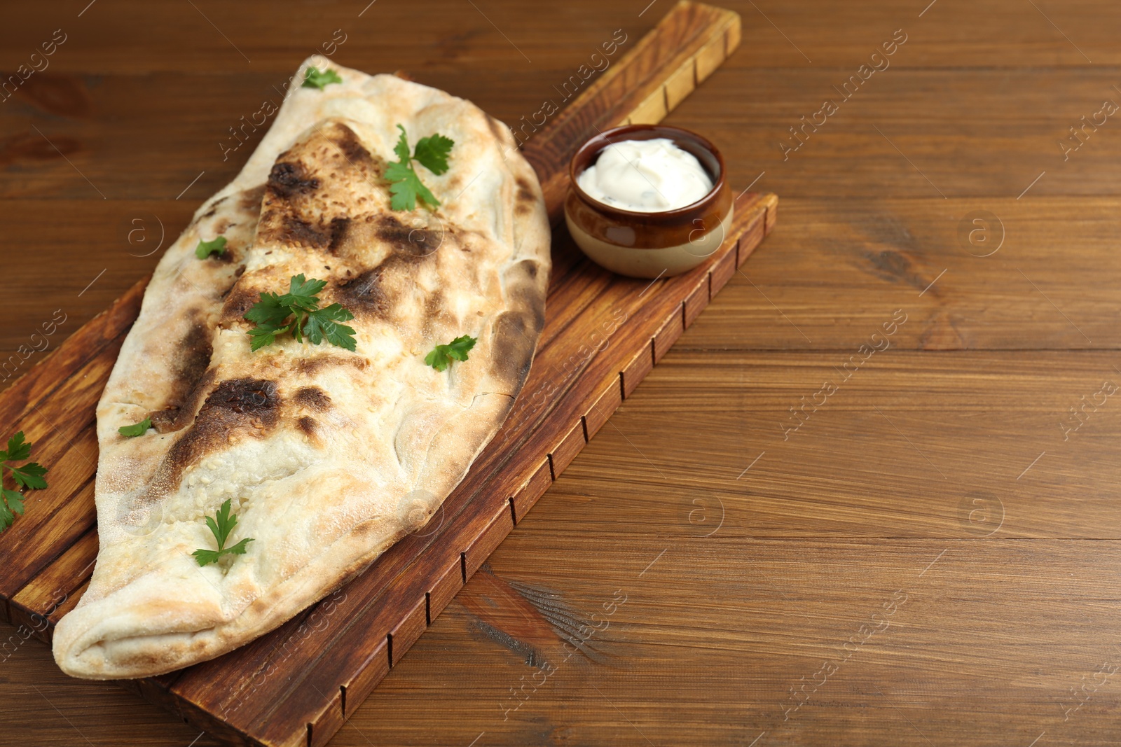 Photo of Board with tasty calzone, parsley and sauce on wooden table, closeup. Space for text