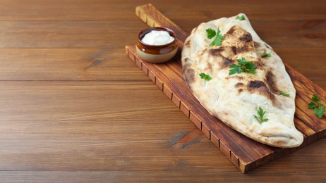 Photo of Board with tasty calzone, parsley and sauce on wooden table, closeup. Space for text