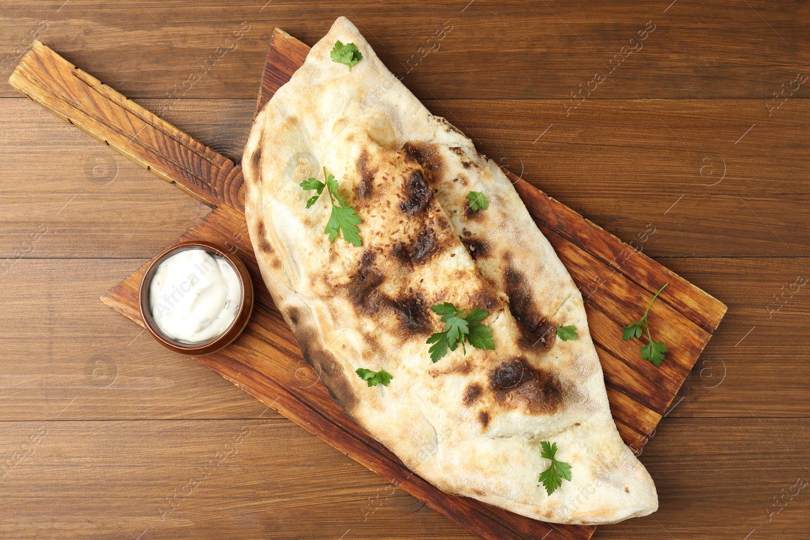 Photo of Board with tasty calzone, parsley and sauce on wooden table, top view