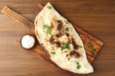 Photo of Board with tasty calzone, parsley and sauce on wooden table, top view