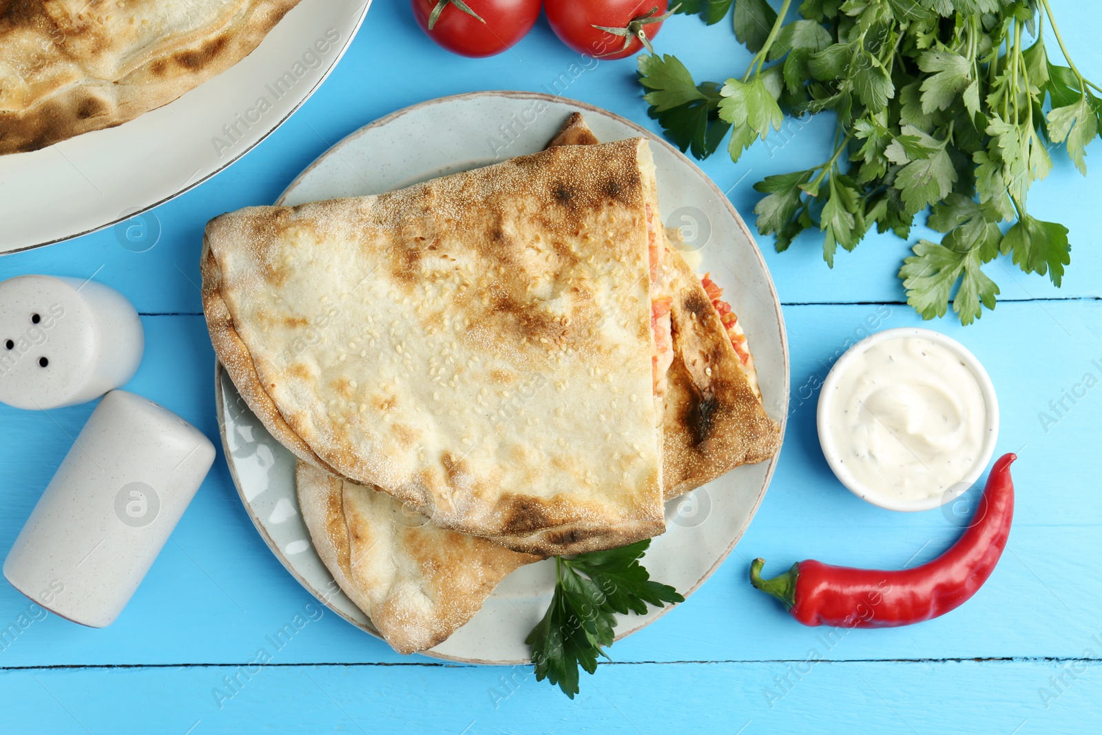 Photo of Halves of tasty calzone, tomato, chili pepper, parsley and sauce on light blue table, flat lay