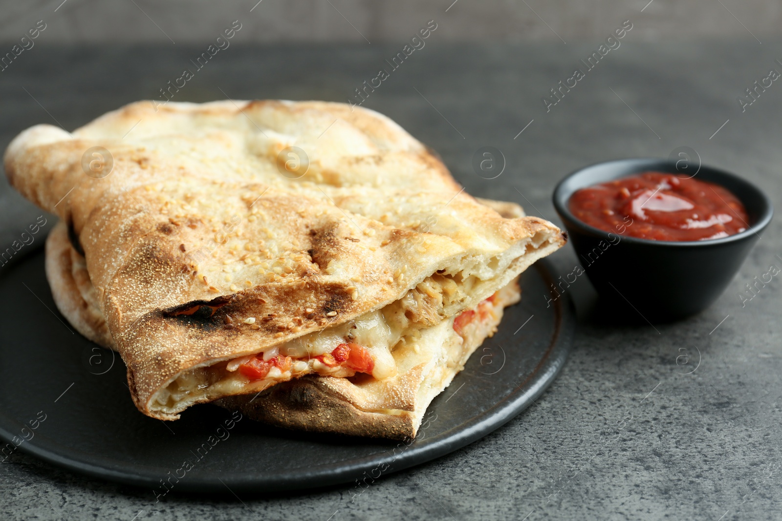 Photo of Halves of tasty calzone with meat, cheese, tomato and sauce on grey textured table, closeup