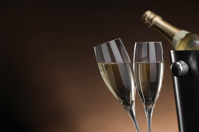 Photo of Champagne in glasses and ice bucket with bottle on brown background, space for text