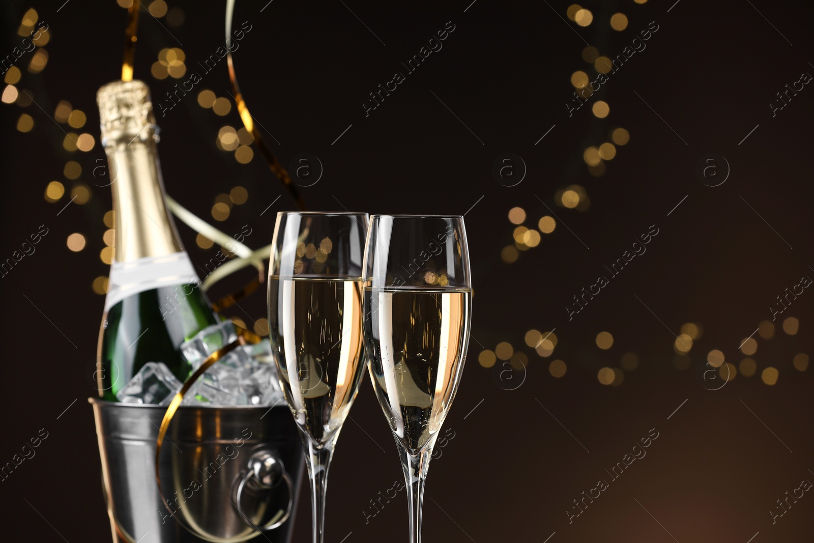 Photo of Champagne in glasses and ice bucket with bottle on brown background with blurred lights, space for text