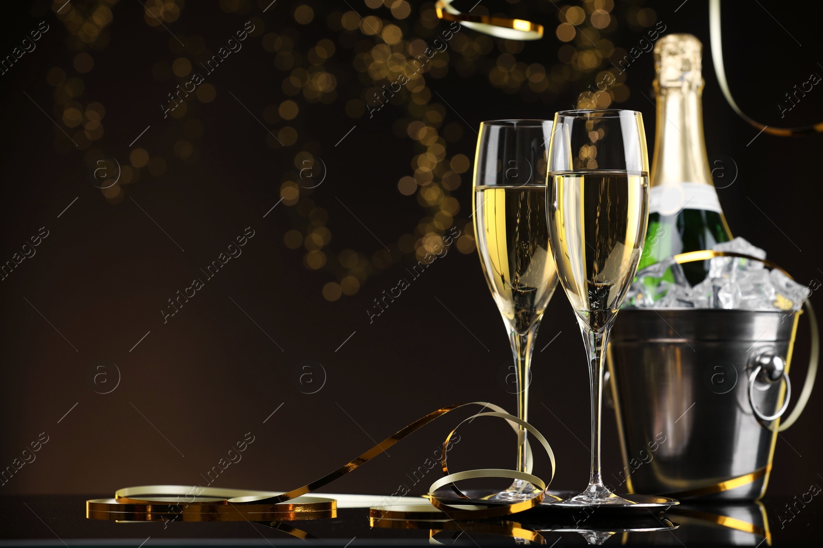 Photo of Champagne and bucket of ice on mirror surface against brown background with blurred lights, space for text