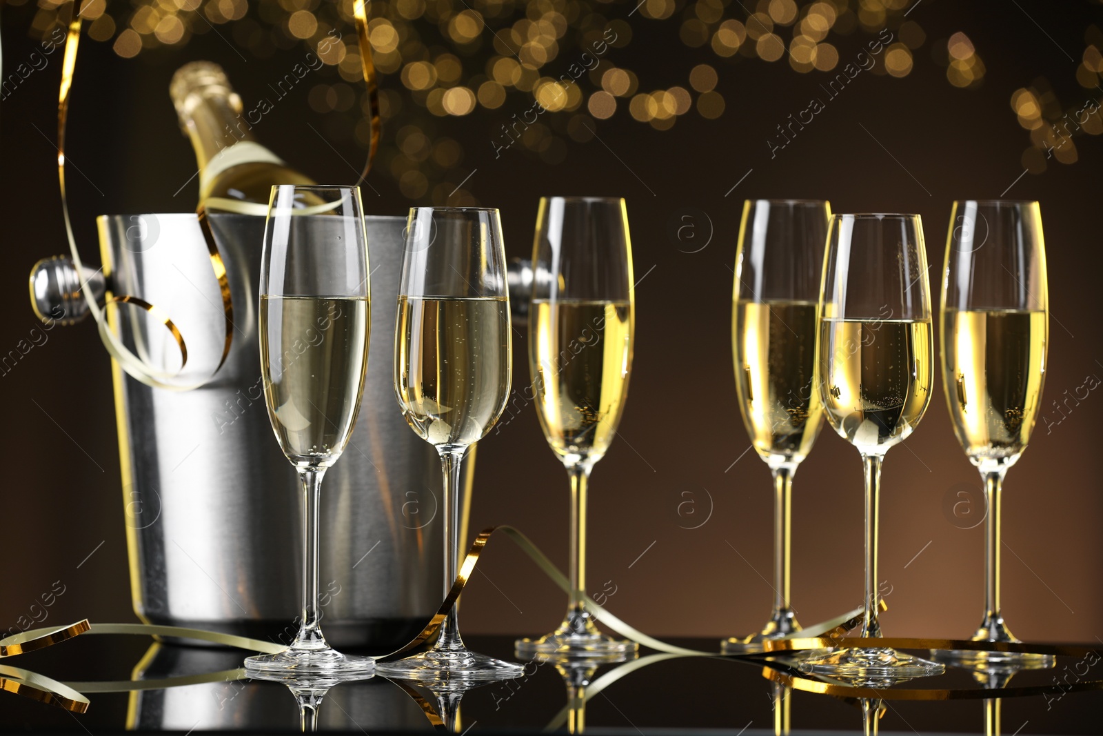 Photo of Champagne in glasses and ice bucket with bottle on mirror surface against brown background with blurred lights