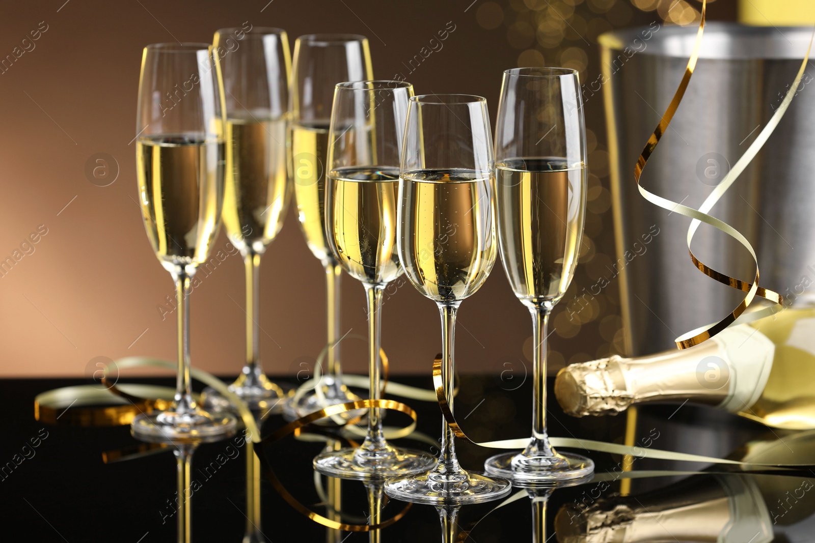 Photo of Champagne in glasses and bottle on mirror surface against brown background with blurred lights