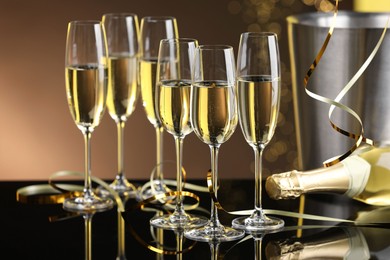 Photo of Champagne in glasses and bottle on mirror surface against brown background with blurred lights