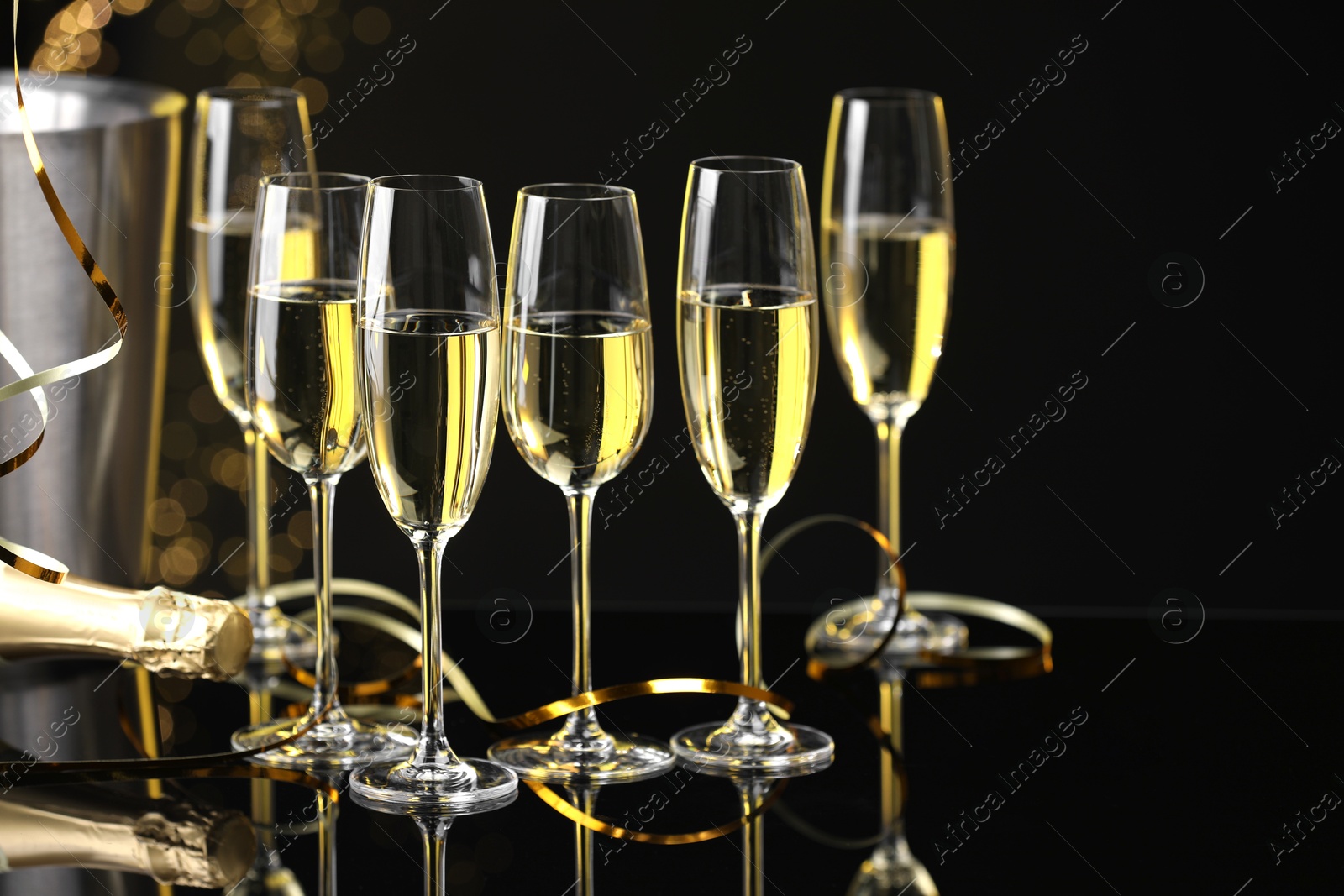 Photo of Champagne in glasses and bottle on mirror surface against black background with blurred lights