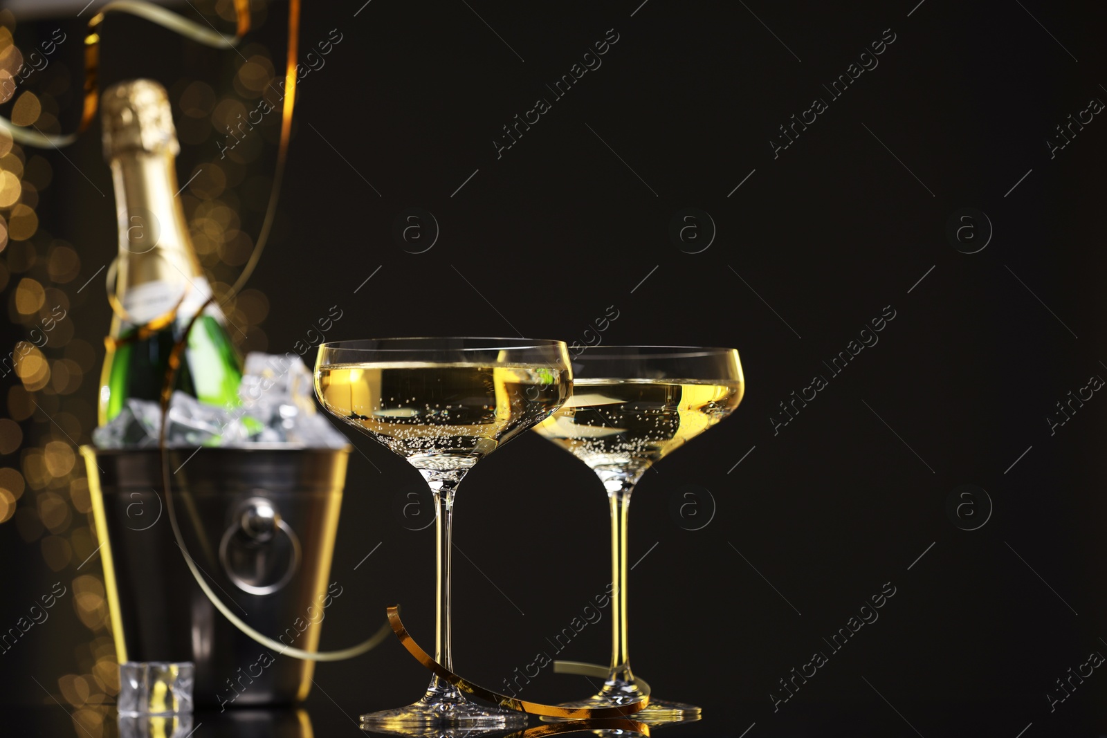 Photo of Champagne in glasses and ice bucket with bottle on mirror surface against black background with blurred lights, space for text