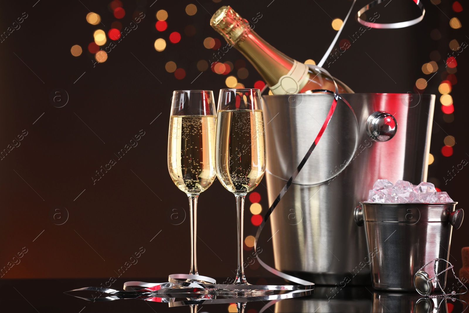 Photo of Champagne in glasses, bottle and ice buckets on mirror surface against color background with blurred lights, space for text