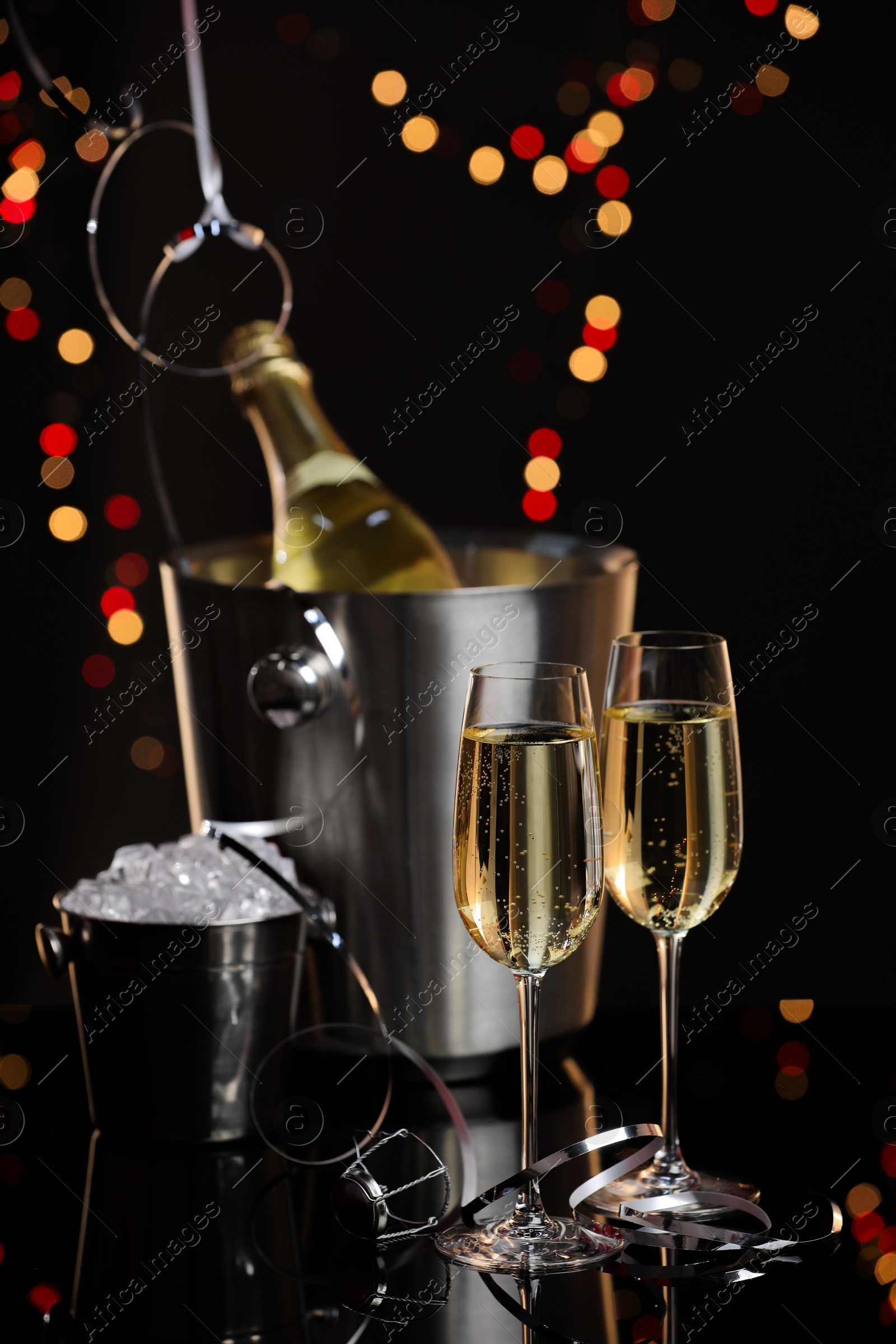 Photo of Champagne in glasses, bottle and ice buckets on mirror surface against black background with blurred lights