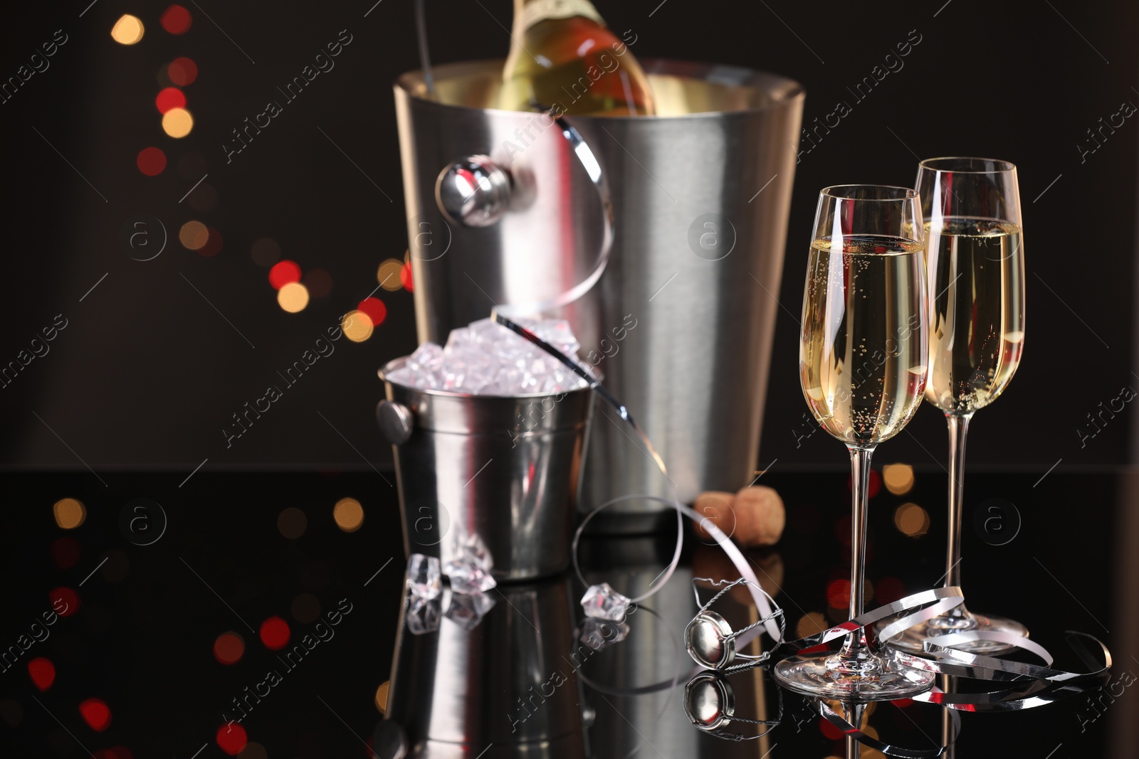 Photo of Champagne in glasses, bottle and ice buckets on mirror surface against black background with blurred lights, space for text