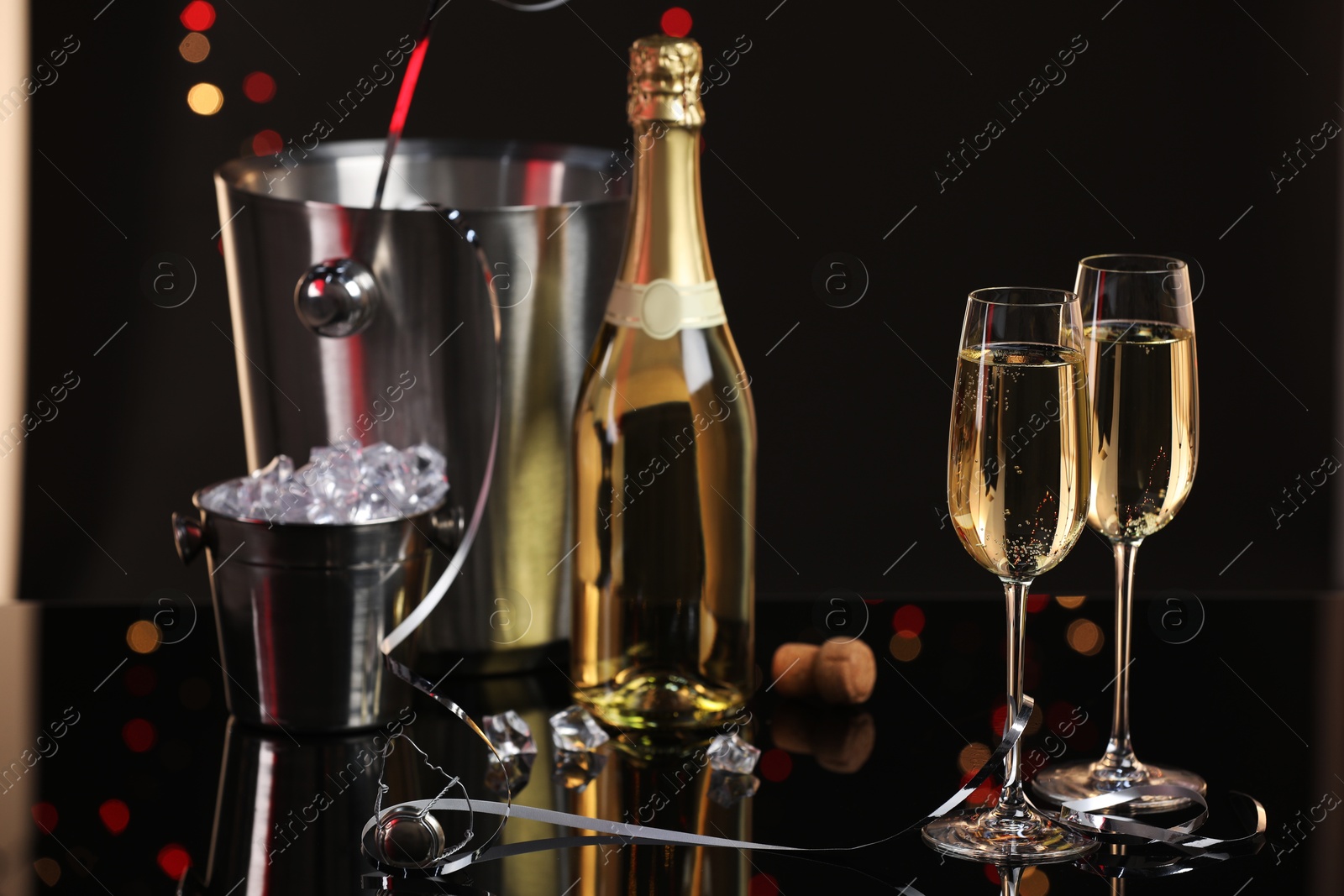 Photo of Champagne in glasses, bottle and ice buckets on mirror surface against black background with blurred lights