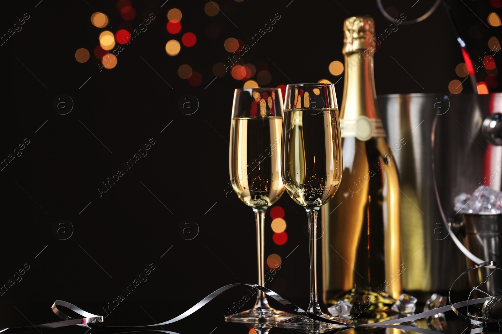 Photo of Champagne in glasses, bottle and ice buckets on mirror surface against black background with blurred lights, space for text