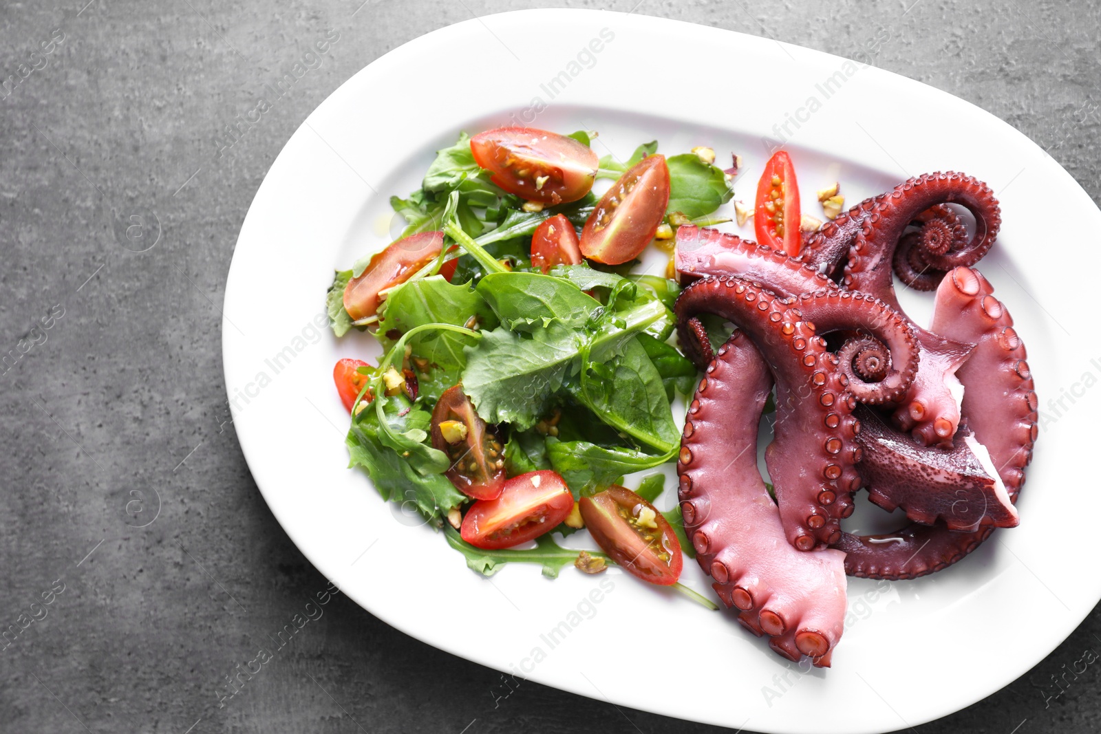 Photo of Plate with tasty boiled octopus tentacles and salad on grey textured table, top view