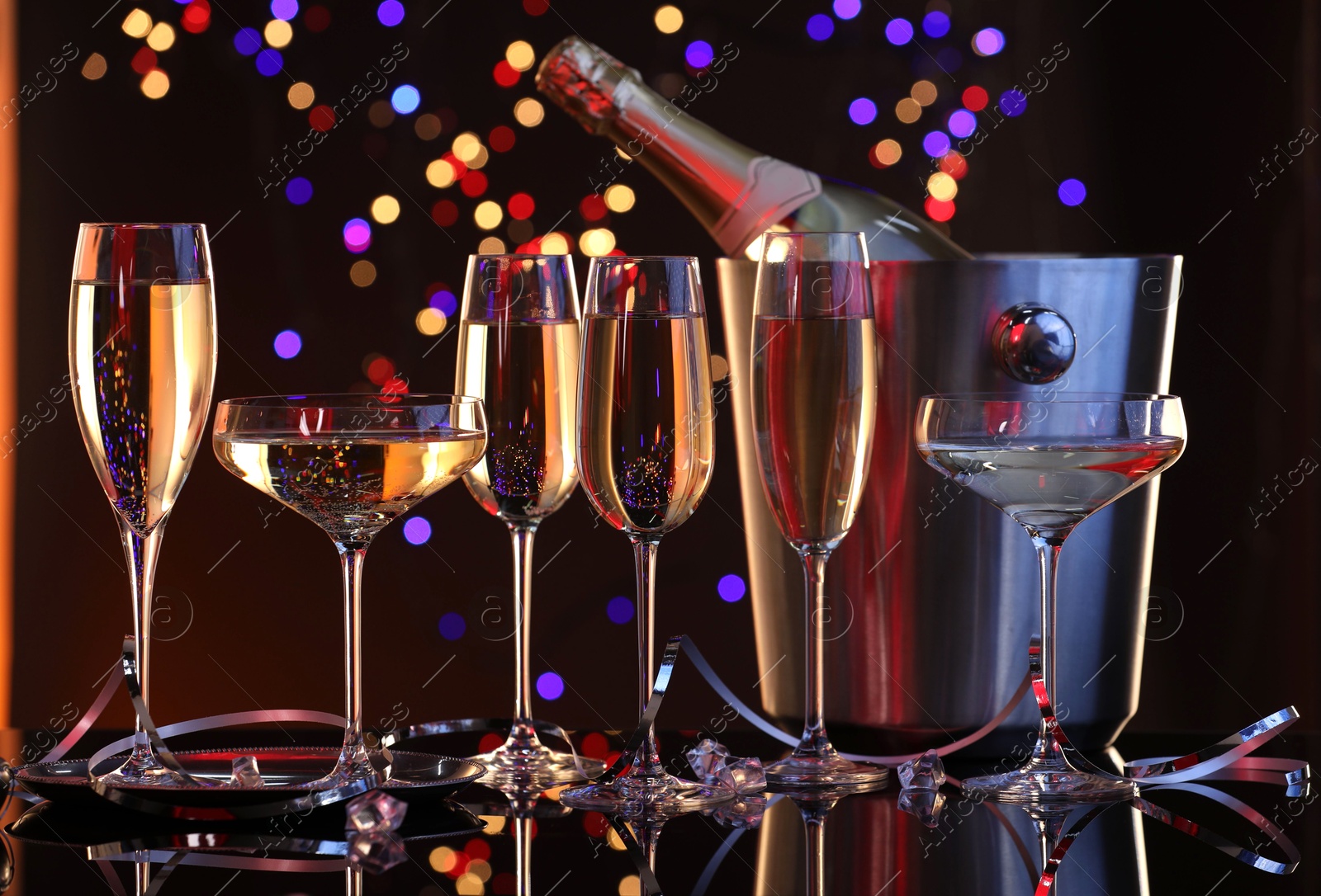 Photo of Champagne in glasses and ice bucket with bottle on mirror surface against dark background with blurred lights