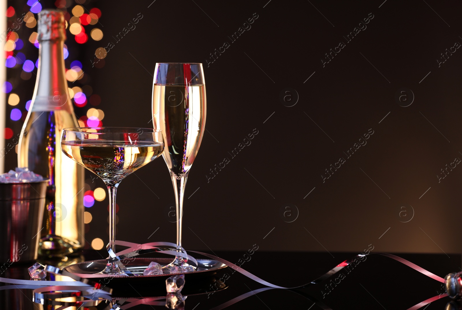 Photo of Champagne in glasses, bottle and ice bucket on mirror surface against brown background with blurred lights, space for text