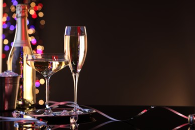 Photo of Champagne in glasses, bottle and ice bucket on mirror surface against brown background with blurred lights, space for text