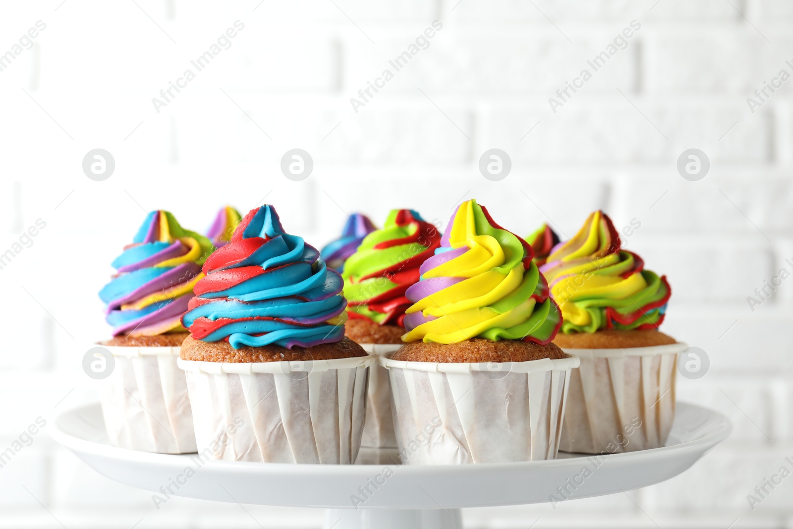 Photo of Tasty cupcakes with colorful cream on stand near white brick wall, closeup