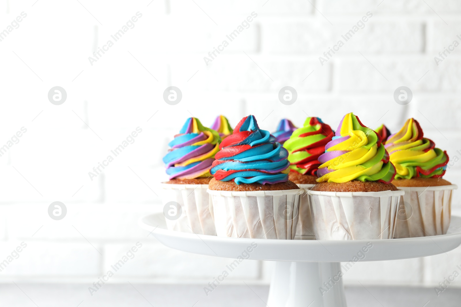 Photo of Tasty cupcakes with colorful cream on stand near white brick wall, closeup. Space for text