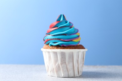 Photo of Tasty cupcake with colorful cream on white table against light blue background, closeup