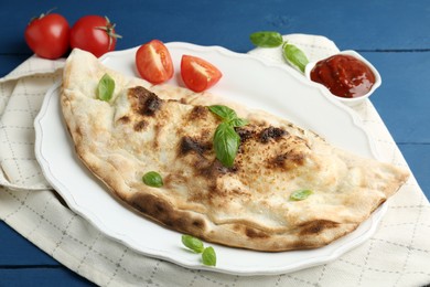 Photo of Tasty calzone with basil, tomatoes and sauce on blue wooden table