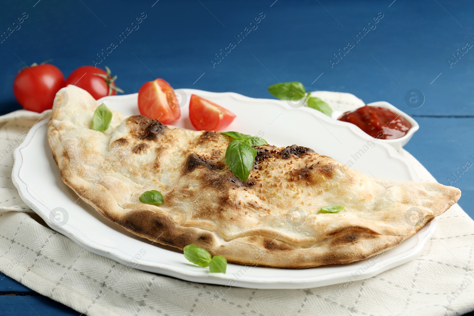 Photo of Tasty calzone with basil, tomatoes and sauce on blue wooden table, closeup