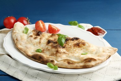 Photo of Tasty calzone with basil, tomatoes and sauce on blue wooden table, closeup