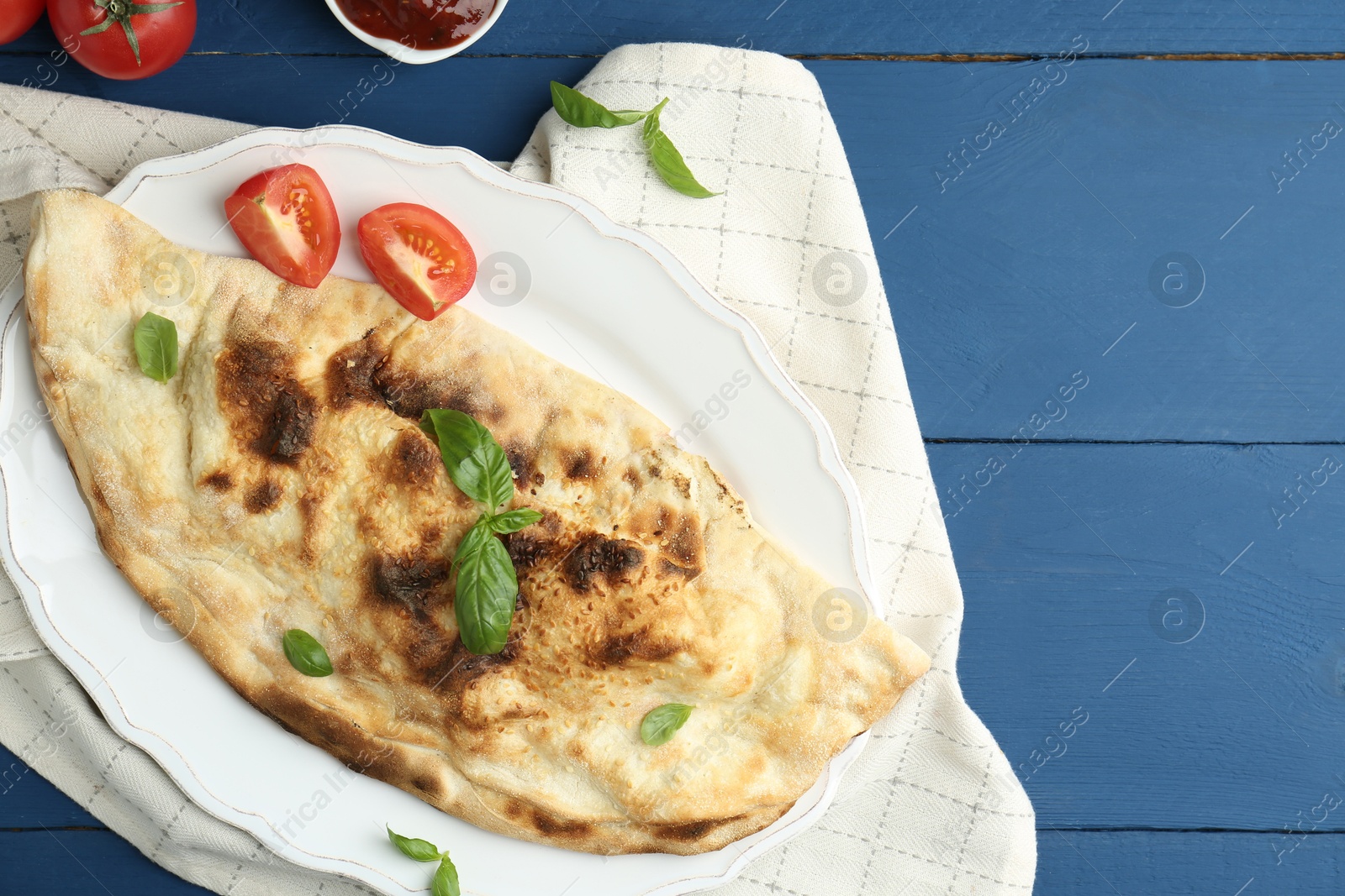 Photo of Tasty calzone with basil, tomatoes and sauce on blue wooden table, flat lay. Space for text